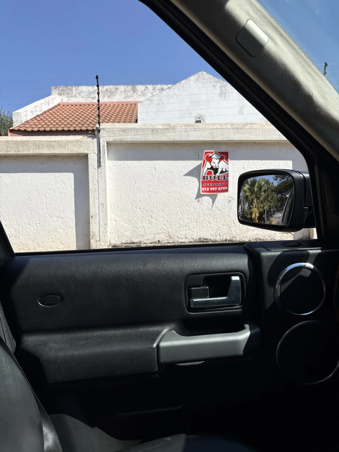 A house and boundary wall photographed through the passenger window of a vehicle.